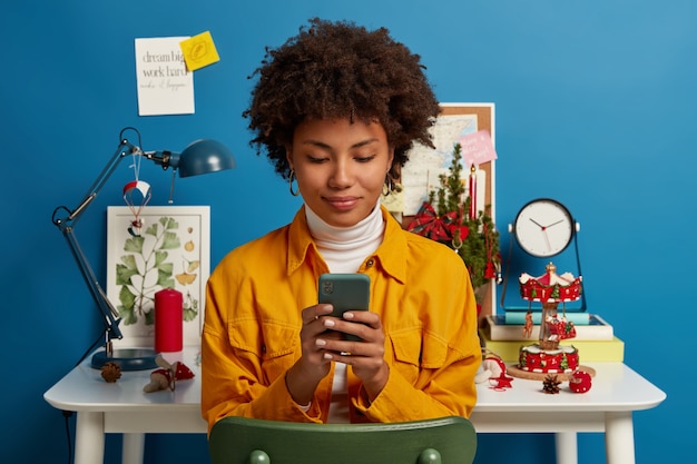 Free photo pleased female student takes break from autodidact, uses cellular for chatting online, browses application, sends text message, checks mail via wifi, sits on chair near workplace, blue wall.