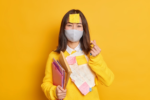Pleased female student wears protective mask to prevent viruses and catching disease has papers with written necessary information stuck on clothes makes korean like sign.