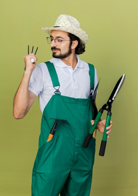 Pleased gardener man in optical glasses wearing gardening hat stands with garden tools