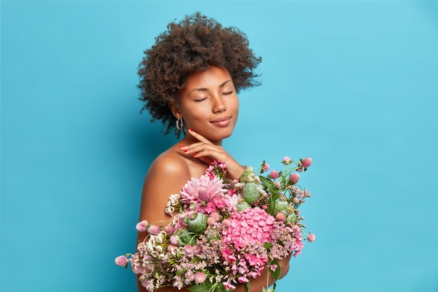 Pleased gentle female model touches jawline closes eyes enjoys lovely moment poses half naked with bunch of beautiful flowers isolated over blue wall