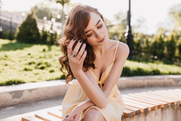 Pleased ginger girl sitting on bench with eyes closed and touching her hair. Outdoor photo of wonderful white woman with curly red hair posing on nature.