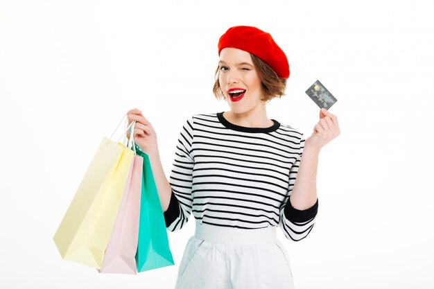 Pleased ginger woman with packages holding credit card and winks at the camera over grey