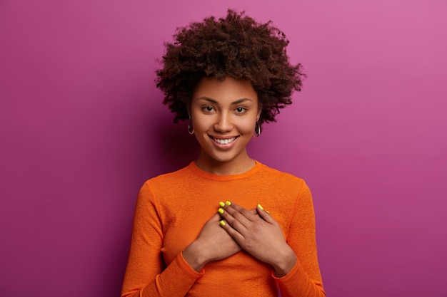 Pleased girl in orange jumper presses palms to heart, makes thankful gesture, touched with cordial congratulations, smiles positively, isolated over purple wall. Acknowledgement concept