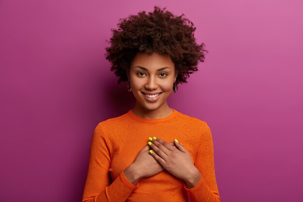 pleased girl in orange jumper presses palms to heart, makes thankful gesture, touched with cordial congratulations, smiles positively, isolated over purple wall. acknowledgement concept
