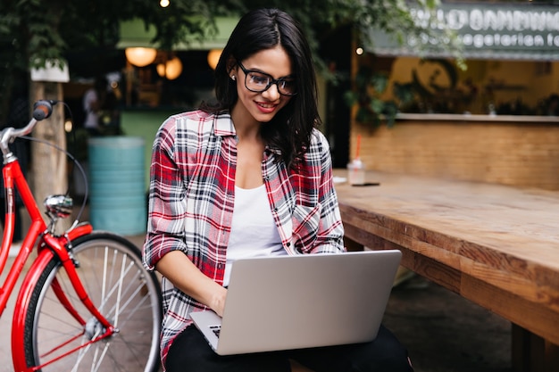 Pleased girl with dark hair looking at laptop screen with smile while sitting on the street. Outdoor photo of interested woman chilling after bike ride.