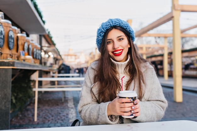 Ragazza soddisfatta con lunghi capelli scuri sorridente per strada mentre beve il caffè. giovane donna sveglia in cappotto e maglione in posa con una tazza di tè in una giornata fredda sulla città.