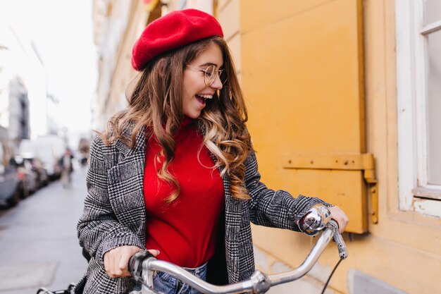 自転車に乗って街の景色を楽しみながら、素敵な巻き毛の女の子を喜ばせます。自転車で週末を過ごすニットベレー帽のロマンチックな若い女性の屋外の肖像画。