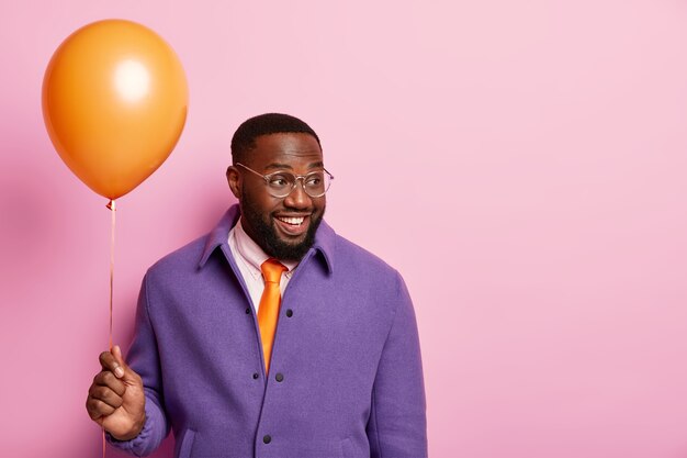 Pleased glad black man with thick beard holds air balloon, has party in office, celebrates promotion, looks happily aside, dressed in festive clothes