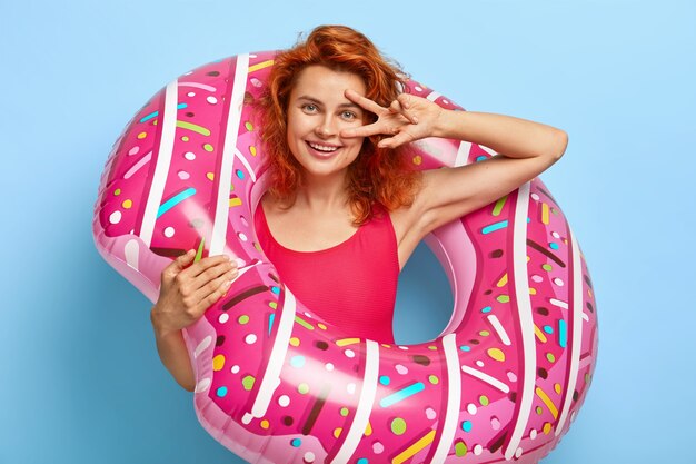 Pleased glad European woman poses with inflatable swim ring, wears fashionable red bikini