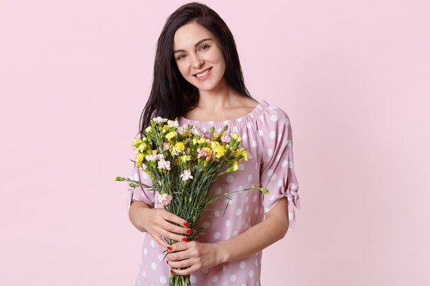 pleased good looking European young woman with gentle smile, wears polka dot dress, holds bouquet of flowers, happy to recieve from husband, models on pink pastel.