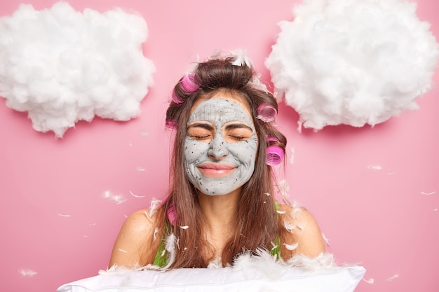 Pleased good looking woman closes eyes smiles positively wears hair rollers on head poses with soft pillow flying feathers around poses against pink wall