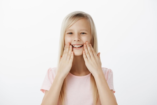 Foto gratuita felice ghignante adorabile bambino con i capelli biondi, sorridente ampiamente e tenendo i palmi vicino alle labbra, stupito e soddisfatto di denti sani, frequentando il dentista e sentendosi felice