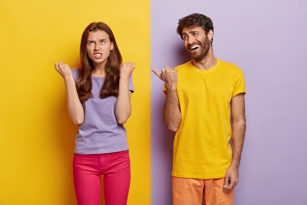 喜んでいる男は楽しんで、明るい黄色のTシャツを着て、怒りで拳を食いしばっているイライラしたガールフレンドに親指を向けます