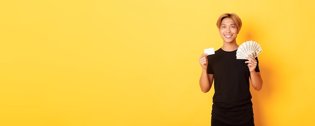 Free photo pleased handsome asian guy showing money and credit card smiling happy standing yellow background