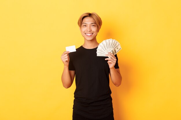 Довольный красивый азиатский парень показывает деньги и кредитную карту, улыбаясь счастливым, стоя желтой стене