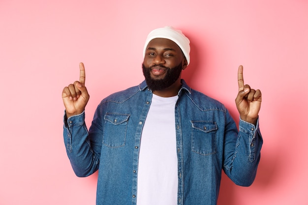 Pleased handsome Black man nodding in approval and checking out good promo, pointing fingers up at offer, showing best deal, pink background.