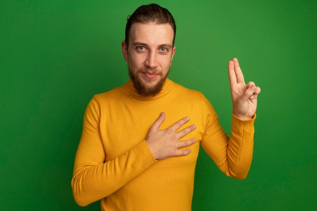 Pleased handsome blonde man doing oath gesture isolated on green wall