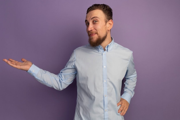 Pleased handsome blonde man holds hand open and looks at front isolated on purple wall