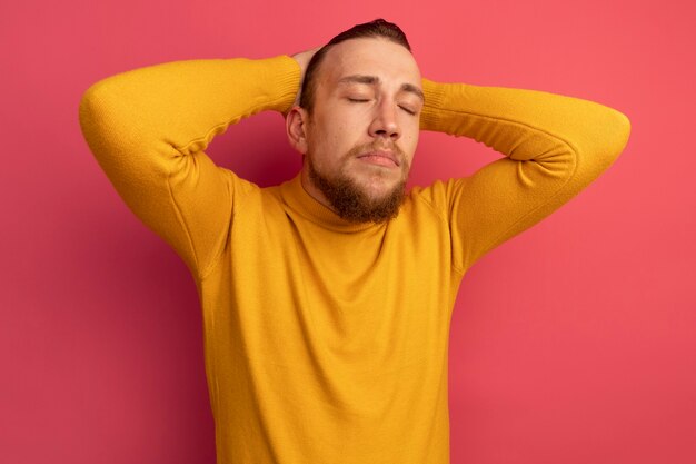 Pleased handsome blonde man stands with closed eyes putting hands on head on pink