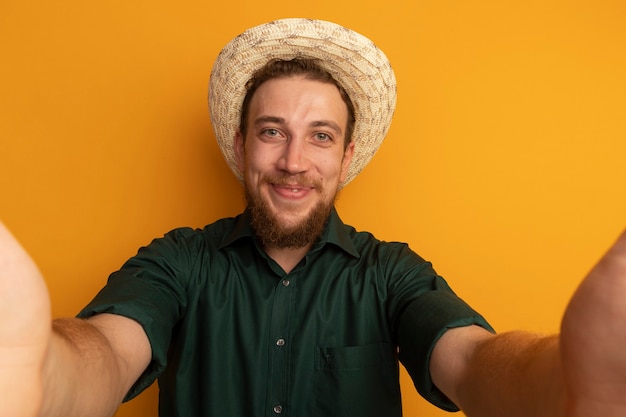 Free photo pleased handsome blonde man with beach hat pretends to hold front isolated on orange wall