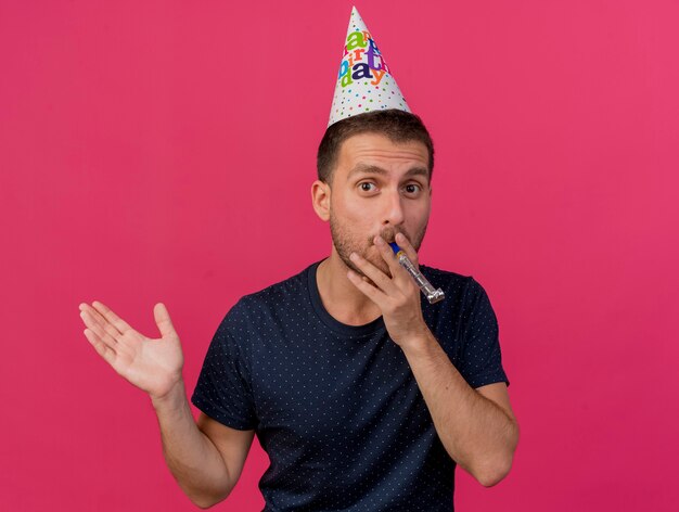 Pleased handsome caucasian man wearing birthday cap blowing whistle isolated on pink background with copy space