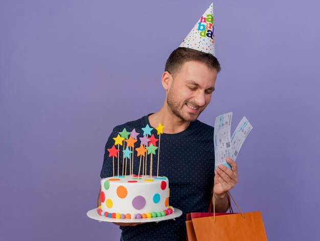 Pleased handsome caucasian man wearing birthday cap holds birthday cake paper shopping bag gift box and air tickets isolated on purple background with copy space