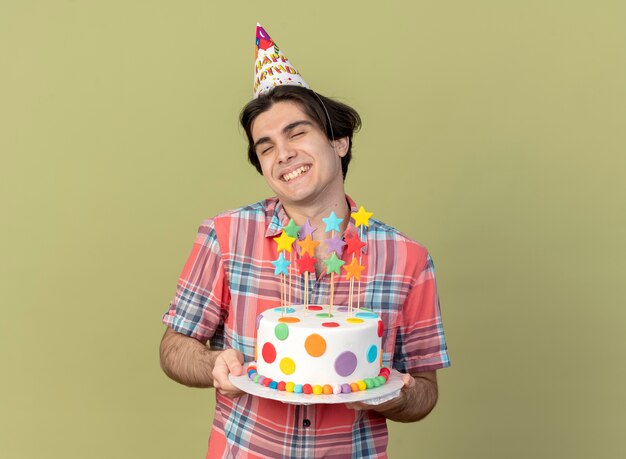 Pleased handsome caucasian man wearing birthday cap holds birthday cake 