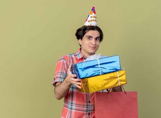생일 모자를 쓰고 기쁘게 잘 생긴 백인 남자는 선물 상자와 종이 쇼핑백을 보유하고 있습니다.
