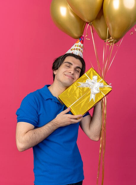 Pleased handsome caucasian man wearing birthday cap holds helium balloons and gift box 