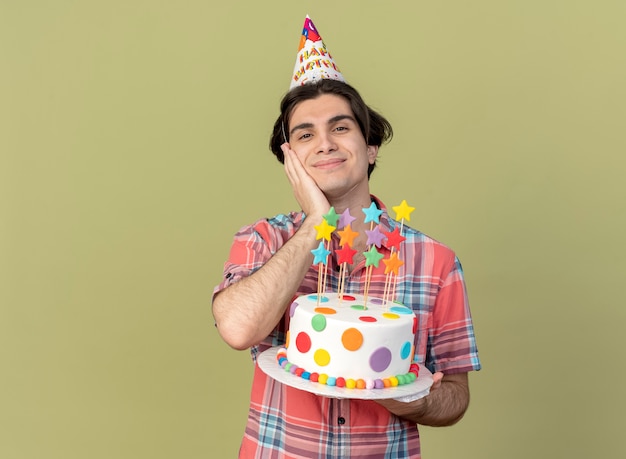 Pleased handsome caucasian man wearing birthday cap puts hand on face and holds birthday cake 