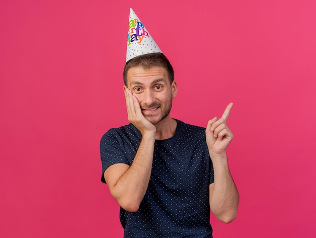 Free photo pleased handsome caucasian man wearing birthday cap puts hand on face pointing at side isolated on pink background with copy space
