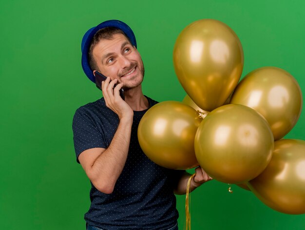 Pleased handsome caucasian man wearing blue party hat holds helium balloons talking on phone looking at side isolated on green background with copy space