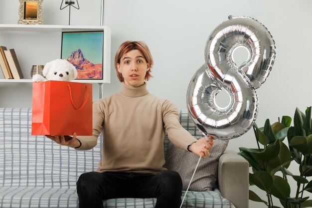 Pleased handsome guy on happy women day holding number eight balloon with gift bag, sitting on sofa in living room