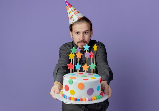 Довольный красавец в кепке на день рождения держит торт ко дню рождения, изолированный на фиолетовой стене