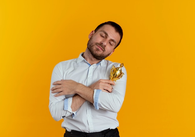 Free photo pleased handsome man holds and hugs winner cup isolated on orange wall