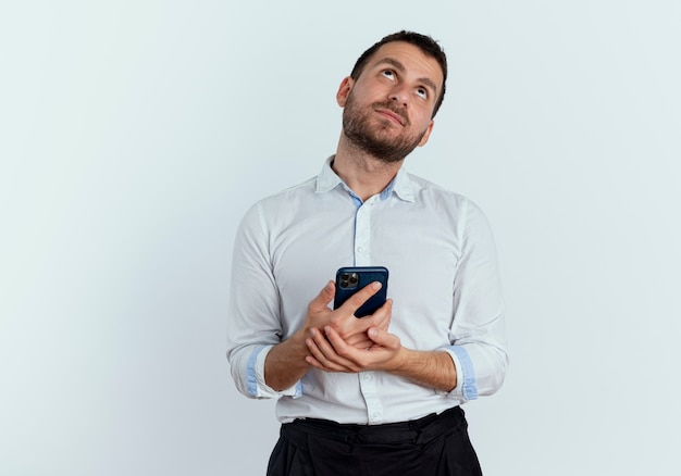 Pleased handsome man holds phone and looks up isolated on white wall