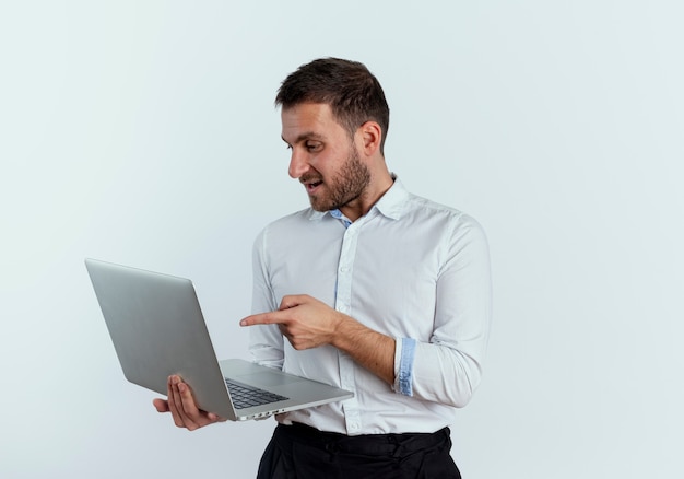 Pleased handsome man looks and points at laptop isolated on white wall