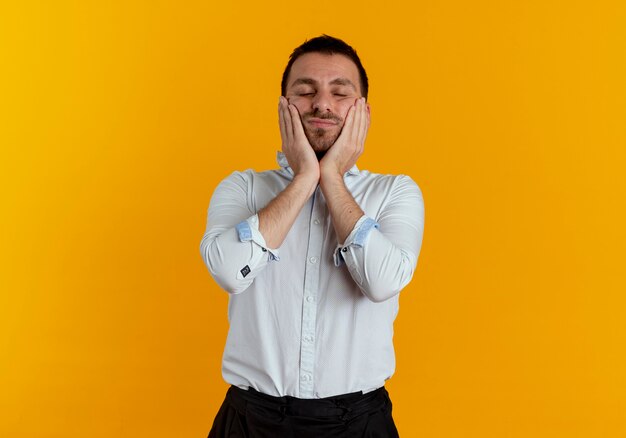 Pleased handsome man puts hands on face isolated on orange wall