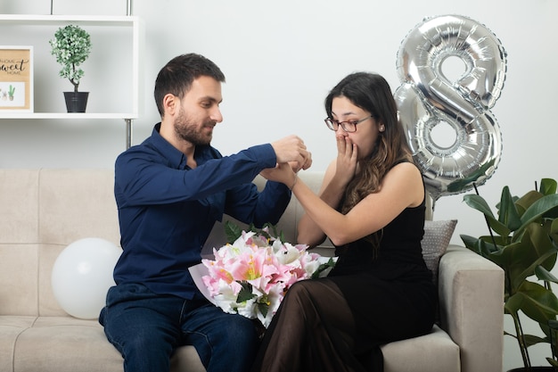 Pleased handsome man putting bracelet on hand of excited pretty young woman in optical glasses holding bouquet of flowers sitting on couch in living room on march international women's day