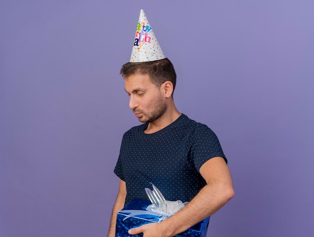 Pleased handsome man wearing birthday cap holds gift box looking at side isolated on purple wall with copy space