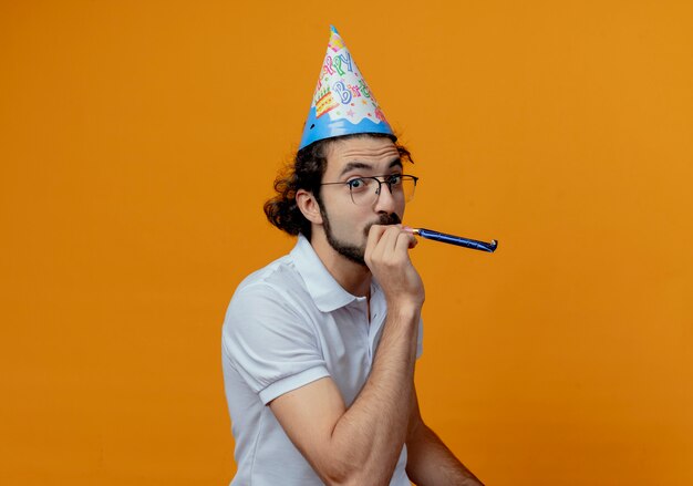 Pleased handsome man wearing glasses and birthday cap blowing whistle  isolated on orange background