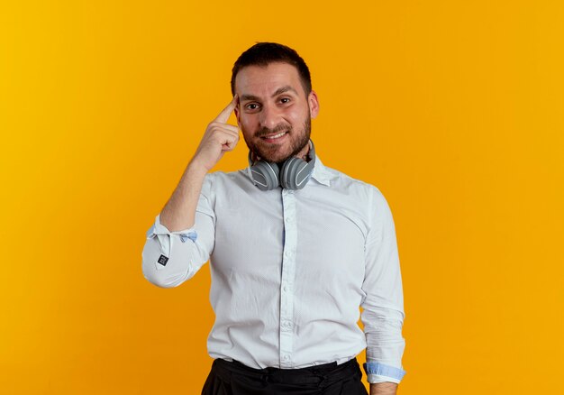 Pleased handsome man with headphones on neck puts finger on temple isolated on orange wall