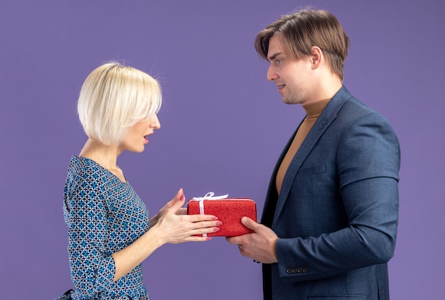 Pleased handsome slavic man giving gift box and looking at surprised pretty blonde woman on valentine's day 