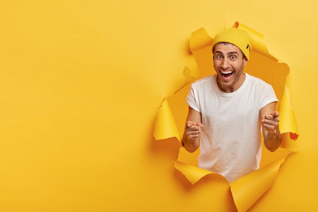 Pleased handsome young man points at you, directs fore fingers at camera, wears white t shirt, yellow headgear, stands in paper hole