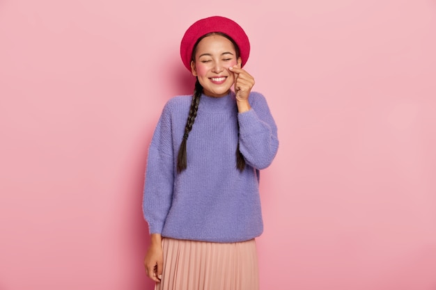 Pleased happy Asian woman shapes small heart with hands, makes korean like sign, wears red beret, casual jumper and skirt, smiles pleasantly, being in good mood, isolated over pink wall