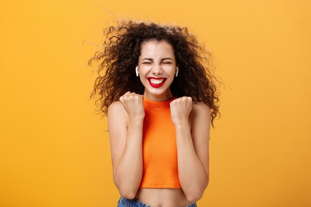 Pleased happy and triumphing curly-haired female in cropped top clenching fists in yes gesture closing eyes and smiling broadly receiving awesome news, winning award over orange background. Copy space