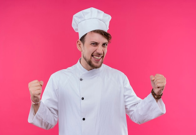 A pleased and happy young bearded chef man in white uniform raising clenched fists in the air while looking on a pink wall