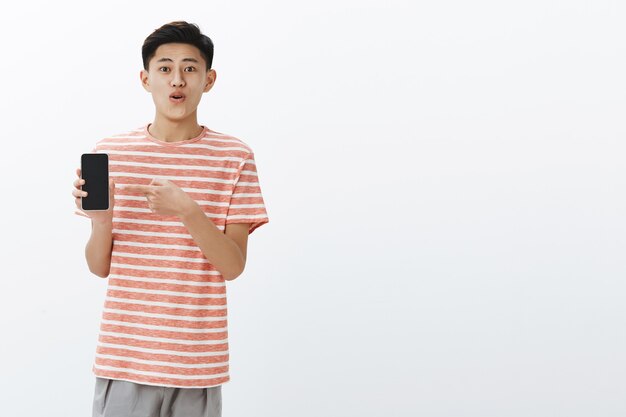 Pleased happy young cute asian guy in striped t-shirt standing to left side of copy space holding smartphone pointing at cellphone screen as showing awesome new phone to friends delighted