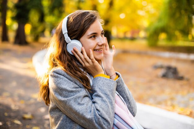 Pleased happy young teenage girl student sitting outdoors in beautiful autumn park listening music with headphones.