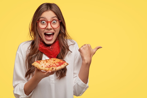 Pleased happy young woman gazes with happiness, points with thumb aside at free space, eats pizza, shows direction, keeps jaw dropped, exclaims in happiness, isolated over yellow wall.